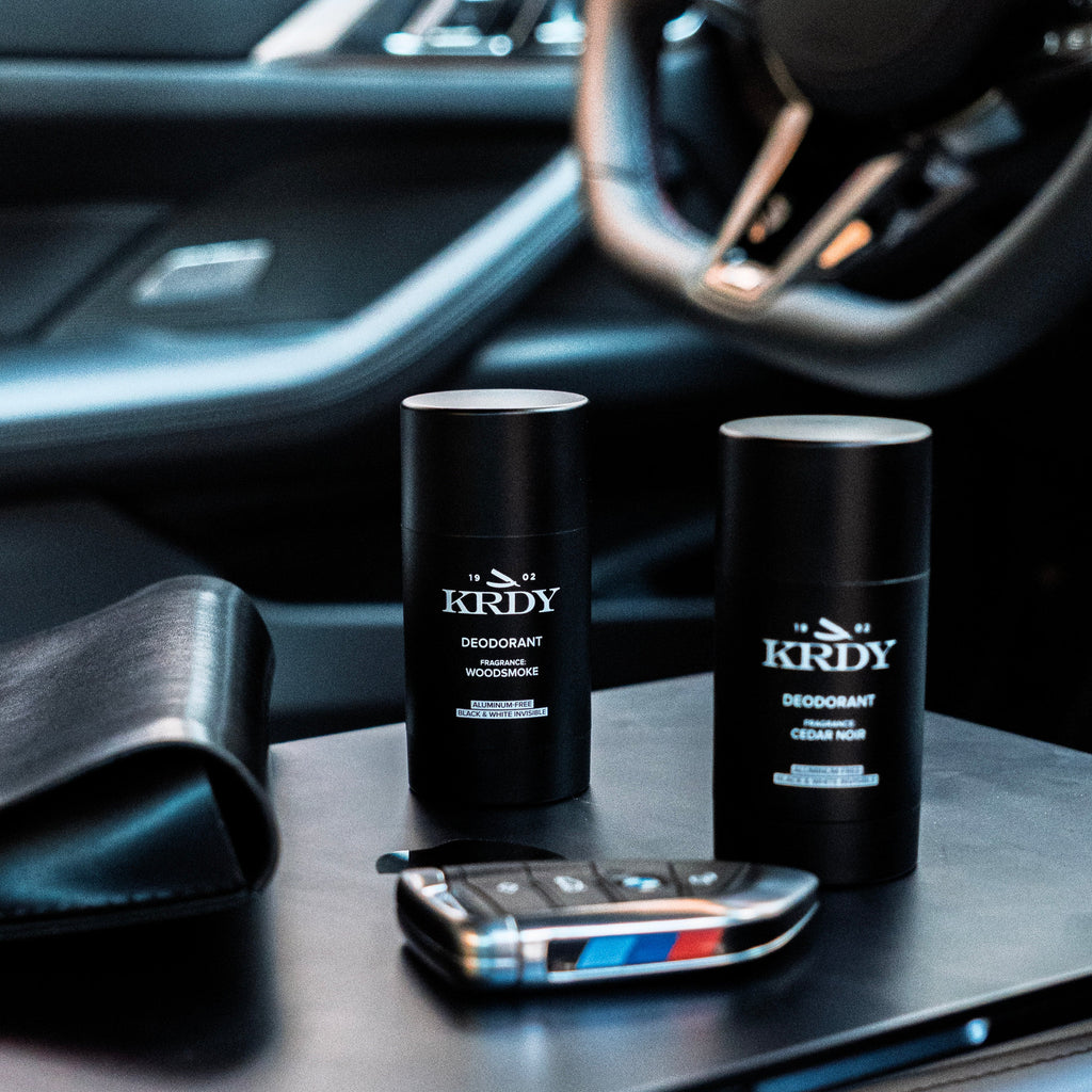 Two KRDY deodorant sticks on a car dashboard with a smartphone and steering wheel in the background.